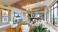 This elegantly designed single-floor communal space presents a sophisticated blend of contemporary and coastal-inspired aesthetics. The interior is immediately distinguished by its striking wood-paneled tray ceilings with recessed lighting and a decorative gold chandelier, which add warmth and architectural character throughout.

The main lounge area is generously proportioned, featuring multiple distinct seating arrangements with plush upholstered sofas, accent chairs in neutral tones of blush, cream, and gray, and sleek mirrored and marble-topped side tables. Large-framed wall art and textured sculptural pieces serve as focal points across the space. A wall-mounted flat-screen television anchors one end of the lounge, complemented by a built-in media console with open shelving.

A dedicated wet bar area is equipped with taupe wood cabinetry, a light quartz countertop, a stainless steel undercounter refrigerator, microwave, and open display shelving with decorative accents. A wall-mounted television above the bar adds functionality to the space.

Natural light flows generously through full-height glass French doors, which open to an outdoor area framed by tropical landscaping. The flooring transitions between a geometric parquet wood pattern and warm area rugs, unifying the space. Lush potted plants are strategically placed throughout, enhancing the bright, resort-like ambiance.