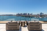 This rooftop terrace offers a striking outdoor experience defined by its generous open-air layout and commanding views of a waterfront harbor and city skyline. The terrace is finished with large-format, light-toned stone tiles that extend across the entire deck, lending a clean and refined aesthetic to the space. Glass panel railings line the perimeter, ensuring unobstructed sightlines across the water and toward the urban skyline beyond, while maintaining a sleek, contemporary appearance.

Multiple seating arrangements are positioned throughout the terrace, featuring dark-toned, weather-resistant outdoor furniture including sofas, lounge chairs, and coffee tables. A partially covered section along one edge of the terrace provides a shaded retreat, with a warm wood-paneled ceiling and recessed lighting overhead. This covered zone adds both architectural interest and functional comfort.

The building's exterior walls, clad in horizontal stone and wood panels, serve as a textural backdrop that complements the minimalist design language of the space. Access to the terrace is provided through a glass-and-aluminum door. The terrace captures abundant natural light throughout the day, making it an ideal outdoor setting for relaxation or social gatherings with exceptional waterfront and skyline scenery.