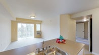 This single-floor residential unit is designed with a functional and comfortable layout. The kitchen is equipped with warm wood cabinetry, a white electric range, a full-size refrigerator, and a double-basin sink set into a laminate countertop that extends into a peninsula, opening directly to the living area. This open-concept arrangement allows natural light from the living room's sliding glass door to filter through the shared space, with outdoor greenery visible beyond. The living area features plush carpeting, a wall-mounted air conditioning unit, and access to what appears to be a balcony or patio.

The bedrooms are carpeted and painted in soft, warm tones, each featuring windows with vertical blinds that allow natural light and outdoor views. Baseboard heaters are present in the bedrooms for supplemental warmth.

The bathroom is well-appointed with a wide wood-toned vanity, ample drawer storage, a large mirror with overhead lighting, and an accessible walk-in shower featuring stainless steel grab bars. Light vinyl flooring completes the clean, practical aesthetic.

A dedicated laundry area houses a full-size washer and dryer, adding everyday convenience. Throughout the unit, the consistent warm palette and practical finishes create a cohesive, livable environment.