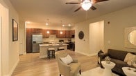 This single-floor residence showcases an open-concept layout that effortlessly connects the kitchen and living area, creating a spacious and functional environment. The kitchen is a standout feature, equipped with rich, dark wood cabinetry that extends to the upper walls, offering generous storage. A central island with light stone countertops provides additional prep space and doubles as a casual dining area, complemented by sleek bar stools. Stainless steel appliances, including a French door refrigerator and range, add a modern, polished touch. Pendant lights hang above the island, contributing to the warm, inviting atmosphere alongside recessed ceiling lighting throughout.

The living area flows naturally from the kitchen, featuring light hardwood flooring that runs consistently across both spaces, enhancing the sense of continuity. A ceiling fan with an integrated light fixture anchors the living area. Neutral, warm-toned walls provide a versatile backdrop, while plush seating and a soft area rug define the lounge space. Decorative wall accents add character without overwhelming the clean, understated aesthetic of the overall design.