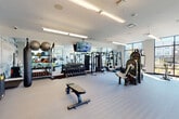 This contemporary fitness center is thoughtfully designed to accommodate a full range of workout activities within a bright, open single-level layout. Large floor-to-ceiling windows with black-framed grid detailing line multiple walls, flooding the space with natural light and providing views of the surrounding outdoor environment. Direct exterior access is available through glass doors, connecting the gym to an outdoor area.

The cardio zone is well-stocked with treadmills, ellipticals, and stationary bikes, all positioned to take advantage of the expansive window views. A dedicated strength training area features a comprehensive dumbbell rack, cable machines, a multi-functional cable cross system, weight benches, and a functional training rig. Additional accessories including resistance bands, stability balls, medicine balls, and a speed bag are neatly organized on wall-mounted racks.

Wall-to-wall mirrors along the strength zone visually expand the space and support proper form during workouts. A bold abstract mural in blue and grey tones adds an artistic focal point. Overhead linear lighting ensures consistent illumination throughout. A water fountain station is also present. The durable, light-toned flooring and clean white walls contribute to a polished, modern aesthetic throughout the facility.