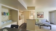 This single-floor unit offers a thoughtfully arranged open-concept layout that seamlessly connects the kitchen, dining, and living areas. The kitchen is outfitted with white cabinetry, a full-size refrigerator, and ample counter space, with a peninsula that serves as a natural divider between the cooking and living zones while maintaining an open, airy feel. The living area features plush carpeting and is furnished with a comfortable sofa and a coffee table, creating a relaxed and inviting atmosphere. Soft, neutral wall tones in warm beige carry throughout the space, contributing to a cohesive and calming ambiance. Decorative artwork adorns the walls, adding a touch of personality to the interior. A standout feature of this unit is the in-unit washer and dryer, conveniently located near the entrance for easy access. Dark hardwood-style flooring in the kitchen and entryway contrasts nicely with the carpeted living space, adding visual interest and defining each functional zone. The layout makes efficient use of space, ensuring each area feels purposeful and well-connected, making this a practical and comfortable single-floor living space.