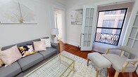 This well-appointed single-floor residence combines classic architectural character with a clean, inviting aesthetic. The living area serves as the heart of the space, showcasing rich hardwood flooring that adds warmth and timelessness to the interior. Crown molding and crisp white trim throughout the room speak to the thoughtful craftsmanship built into the design.

A standout feature of the living area is the full-length French door with louvered panels that opens to a balcony, allowing natural light to pour into the space while offering views of the surrounding brick streetscape — a detail that lends the residence an urban, classic charm. The neutral, light-toned walls create a versatile backdrop that enhances the sense of brightness and openness.

The layout flows naturally from the living area into additional rooms, as seen through the open doorway leading deeper into the residence, suggesting a well-connected and practical floor plan. The space is designed to maximize both comfort and functionality, with proportions that feel welcoming without being overwhelming.

Overall, the residence carries a refined yet approachable ambiance — a blend of traditional architectural details and light, airy interiors that create a comfortable and stylish place to call home.