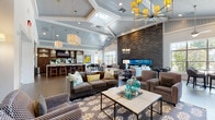 Located at 1138 Fawn Dr in Williamstown, NJ, this single-floor clubhouse is defined by its striking architectural design and carefully curated interior. The centerpiece of the main lounge is a floor-to-ceiling stacked stone accent wall housing a wide linear fireplace, flanked by symmetrical seating arrangements with upholstered sofas, accent chairs, and patterned area rugs. Soaring vaulted ceilings with painted tongue-and-groove paneling in soft blue-gray tones lend a coastal elegance to the space, while an assortment of pendant chandeliers and statement light fixtures provide warm, layered illumination throughout.

Dark wood-look tile flooring flows seamlessly across the open layout, connecting the lounge areas to a well-appointed kitchen and bar. The kitchen features rich dark cabinetry, a long white quartz island with bar stool seating, stainless steel appliances, and industrial-style pendant lighting. Illuminated glass shelving and textured wall tile add refined detail.

An additional seating area with a built-in media center and display cabinetry offers a more intimate gathering space, while a small café-style table near a large grid window provides a bright spot with outdoor views. Decorative wall art and carefully placed accents complete the polished, contemporary ambiance throughout.