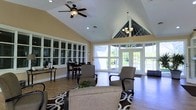Located at 2979 Green Arbor Lane in Dublin, Ohio, this single-floor community space offers a warm and inviting atmosphere designed for comfort and social engagement. The interior is anchored by an impressive vaulted ceiling that rises dramatically overhead, accented by a classic ceiling fan and an elegant chandelier suspended beneath a striking pyramid-style skylight structure that floods the space with natural light. Large glass French doors along the rear wall open to an outdoor view of lush greenery, seamlessly blending the interior with the surrounding landscape. The flooring throughout features rich hardwood tones that complement the neutral warm-beige walls and crisp white trim. The lounge area is furnished with plush upholstered seating arranged on a boldly patterned area rug, creating a comfortable gathering space. Adjacent to the lounge, a casual dining area provides additional seating with bar-height tables and chairs, ideal for informal meals or social gatherings. A built-in cabinetry wall with glass-paned doors adds a refined architectural detail and practical storage. Recessed lighting and ample windows ensure the space remains bright throughout the day. Overall, the layout strikes a balance between open, airy design and purposeful functionality.
