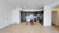 This single-floor space showcases a thoughtfully designed open-concept layout that seamlessly integrates the kitchen and living areas. The kitchen is a standout feature, equipped with full-height dark espresso cabinetry that provides generous storage from floor to ceiling. A classic white subway tile backsplash adds a clean, timeless contrast against the dark cabinetry, while a suite of stainless steel appliances — including a French door refrigerator, electric range, and over-the-range microwave — delivers a sleek, modern functionality. A freestanding wooden kitchen island with a lower storage shelf serves as a practical and versatile workspace, ideal for meal preparation or casual dining. The flooring throughout is light-toned, wood-style plank, which contributes warmth and continuity across the open space. Recessed lighting in the kitchen area ensures bright, even illumination. The open floor plan creates a sense of spaciousness and ease of movement, making the layout feel airy and connected. The neutral white walls and clean architectural lines lend a contemporary, minimalist aesthetic that is both inviting and functional.