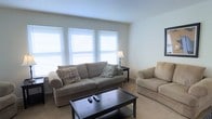 This two-story space at 3801 Veteren offers a clean, functional layout with a neutral and welcoming aesthetic throughout. The living area, as seen, is bathed in natural light streaming through a trio of large windows fitted with white horizontal blinds, creating a bright and airy atmosphere. The room features light-colored carpeting that complements the soft, beige-toned upholstered seating, including a full sofa accented with decorative throw pillows and a matching loveseat. A dark-finished coffee table serves as a central anchor for the seating arrangement, adding a subtle contrast to the room's otherwise light palette. A classic table lamp provides supplemental ambient lighting. The walls are painted in a crisp, clean white, contributing to the open and spacious feel of the living area. The two-floor layout of this space provides distinct zones for living and other activities, offering a functional separation between levels. Overall, the space presents a straightforward, well-lit environment with a simple, comfortable design sensibility suited for everyday living.
