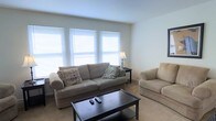 This two-story space at 3801 Veteren offers a clean, functional layout with a neutral and welcoming aesthetic throughout. The living area, as seen, is bathed in natural light streaming through a trio of large windows fitted with white horizontal blinds, creating a bright and airy atmosphere. The room features light-colored carpeting that complements the soft, beige-toned upholstered seating, including a full sofa accented with decorative throw pillows and a matching loveseat. A dark-finished coffee table serves as a central anchor for the seating arrangement, adding a subtle contrast to the room's otherwise light palette. A classic table lamp provides supplemental ambient lighting. The walls are painted in a crisp, clean white, contributing to the open and spacious feel of the living area. The two-floor layout of this space provides distinct zones for living and other activities, offering a functional separation between levels. Overall, the space presents a straightforward, well-lit environment with a simple, comfortable design sensibility suited for everyday living.