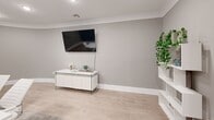 Located at 175 Quincy Ct in Perth Amboy, NJ, this polished single-room space offers a refined, contemporary environment ideal for professional meetings and gatherings. The room is defined by a striking architectural curved wall, finished in a warm neutral gray and adorned with a sculptural metallic wall piece, lending the space a distinctive and sophisticated character.

Light wood-look flooring runs throughout, complementing the soft gray walls and crisp white crown molding that frames the ceiling. A bold accent wall in a deep slate blue adds visual depth and contrast, accented by a framed abstract artwork. Recessed lighting and a modern pendant fixture provide ample illumination across the space.

At the center of the room, a sleek white conference table is surrounded by contemporary white upholstered chairs, anchored by a gray area rug beneath. A wall-mounted flat-screen television is positioned above a modern white credenza with glass legs, which serves as a functional media and display surface. A geometric white open shelving unit along one wall showcases curated decorative accents and lush trailing greenery, adding warmth to the otherwise clean-lined design.

The space is accessed through a glass-paneled door, maintaining an open and transparent feel. Overall, the room delivers a bright, professional, and elegantly appointed setting.