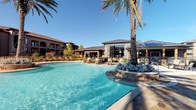 This outdoor pool area offers a well-appointed retreat designed for relaxation and recreation. The centerpiece is a generously sized swimming pool with crystal-clear blue water, framed by a broad concrete sun deck lined with lounge chairs and shade umbrellas — ideal for sunbathing or socializing. Tall palm trees are strategically placed throughout the space, adding a resort-inspired ambiance and natural shade. A dedicated pool house structure serves the area, featuring stucco exterior walls, dark wood-framed glass doors, and direct sightlines to the pool through large windows. The pool house interior provides a transitional space between the building and the pool deck. A separate spa or hot tub is also present, positioned adjacent to the main pool for convenient access. The surrounding multi-story residential buildings frame the space on multiple sides, creating a semi-enclosed, community-oriented atmosphere. Lush landscaping with native plantings and decorative stone accents add texture and visual interest throughout. The overall design balances practicality with aesthetic appeal, offering a comfortable and inviting outdoor amenity space suited for everyday use.