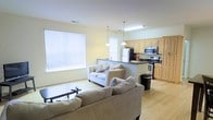 This single-floor space features an open-concept layout that connects the living area and kitchen in a cohesive, functional design. The living room is finished with light hardwood flooring and pale yellow walls, creating a warm, airy atmosphere. Natural light flows generously through large windows fitted with white horizontal blinds, further brightening the space. The living area comfortably accommodates multiple seating pieces, including neutral-toned upholstered sofas accented with decorative pillows.

The kitchen is separated from the living area by a raised counter peninsula with a dark contrasting countertop, providing both a visual boundary and additional prep or seating space. Light maple cabinetry lines the kitchen walls, offering ample storage, while stainless steel appliances — including a refrigerator, microwave, and range — contribute a modern, practical touch. Two pendant lights hang above the peninsula, adding focused task lighting that complements the overhead fluorescent fixture.

The overall ambiance is clean, bright, and contemporary, with consistent flooring and a neutral color palette that unifies the open layout into a cohesive, welcoming living environment.