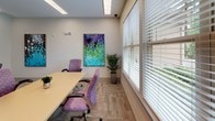 Located at 350 Bradley Park Ln in Cumming, Georgia, this single-floor office space is designed to support a productive and professional work environment. The layout includes a dedicated conference area furnished with a light wood table and ergonomic purple upholstered chairs, ideal for meetings and collaboration. Large windows fitted with horizontal blinds flood the space with natural light while offering views of the surrounding greenery, creating a bright and welcoming atmosphere. Interior glass panels and a glass-paned door connect the conference room to adjacent areas, promoting an open and transparent feel throughout the office. A separate workstation area features two built-in desks equipped with desktop computers, a multifunction printer, and ample cabinetry for storage. The flooring throughout consists of neutral, patterned carpet tiles that complement the light-toned walls. Decorative touches, including vibrant abstract artwork and potted tropical plants, add color and life to the space. Recessed and panel lighting ensures consistent illumination across all areas. This well-organized office space offers a balance of functionality and comfort, making it well-suited for both focused individual work and group collaboration.