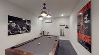 The billiards room at The Lofts at Middlesex is a well-appointed amenity space designed for recreation and socializing. The centerpiece of the room is a full-size pool table with a gray felt surface and rich wood finish, positioned beneath a striking cluster pendant light fixture with industrial-style metal dome shades that cast focused illumination across the playing surface. Recessed ceiling lights supplement the overhead lighting, ensuring the room remains bright and functional throughout.

The walls are finished in crisp white, providing a clean, modern backdrop that highlights the room's carefully chosen décor. A bold movie poster for "The Color of Money" adds a thematic touch, nodding to the classic billiards film, while a black-and-white photographic print of players around a pool table reinforces the room's dedicated purpose. A wall-mounted cue rack keeps several cue sticks and a triangle rack neatly organized and easily accessible.

Dark gray carpet covers the floor, adding a layer of comfort underfoot while complementing the room's contemporary aesthetic. A compact high-top table with two red adjustable bar stools provides a casual seating area for spectators or those waiting their turn. A wall-mounted mini-split climate control unit ensures year-round comfort. The overall ambiance is modern, focused, and inviting.