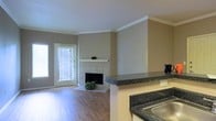 This single-floor residence offers a well-designed open-concept layout that seamlessly connects the living and kitchen areas. The living room is a welcoming space featuring warm hardwood flooring, neutral taupe walls, and a classic tiled fireplace with a white mantel that serves as a natural focal point. Crown molding along the ceiling adds a refined architectural touch throughout. A door with slatted blinds allows natural light to filter into the living area, creating a bright and airy atmosphere. The kitchen is visible from the living space, separated by a half-wall breakfast bar topped with dark granite-style countertops that provide both function and style. The sink area and ample counter space make the kitchen practical for everyday use. The open sightlines between the kitchen and living room enhance the sense of spaciousness, making the layout ideal for both daily living and entertaining. The consistent neutral color palette and coordinated finishes throughout give the space a cohesive, move-in-ready feel. Overall, this residence combines classic design elements with functional living spaces on a single, easy-to-navigate floor.