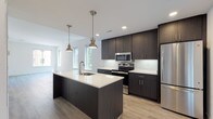 This single-floor unit showcases a contemporary design aesthetic with clean lines and a neutral palette throughout. The open-concept living and kitchen area serves as the heart of the space, where light hardwood flooring extends seamlessly from the living area to the kitchen. Large windows along the living room wall invite abundant natural light, creating a bright and airy atmosphere.

The kitchen is a standout feature, equipped with dark espresso-toned cabinetry that reaches toward the ceiling for ample storage. A central island with a white quartz countertop and an integrated sink serves as both a workspace and gathering point, topped with two stylish brushed-nickel pendant lights. Stainless steel appliances, including a refrigerator, range, and over-the-range microwave, complete the modern culinary space.

The bedrooms feature plush carpeting and generous natural light from multiple windows with urban views. One bedroom offers access to a private balcony through sliding glass doors. Both bathrooms mirror the kitchen's design language, featuring dark wood-grain vanities, white quartz countertops, large mirrors, and neutral-toned flooring, providing a cohesive and polished finish throughout the unit.