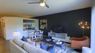 This single-floor space showcases a thoughtfully designed open-concept layout that combines the living, dining, and kitchen areas into one cohesive environment. The living room is anchored by a striking dark accent wall that creates a dramatic focal point, complemented by a flat-screen television mounted on a sleek, light-toned media console. A generously sized sofa in neutral tones is paired with blue accent pillows and a tufted leather ottoman, layered over a patterned area rug that adds warmth and texture to the space. An overhead ceiling fan with an integrated light fixture provides both comfort and ambient lighting. Adjacent to the living area, a compact dining nook features a small table with white chairs, ideal for everyday meals. The kitchen is visible in the background through an open pass-through or shared sightline, offering a bright contrast to the moody tones of the living space. Abstract wall art adds a curated, contemporary touch, while the overall color palette — combining deep charcoal, warm neutrals, and blue accents — gives the space a modern yet inviting ambiance throughout.