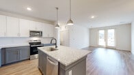 Located at 3355 Club Dr in Lawrenceville, GA, this single-floor home offers a thoughtfully designed open-concept layout that seamlessly connects the kitchen and living areas. The kitchen is a standout feature, showcasing a large center island topped with light granite countertops, an undermount sink with a matte black faucet, and stylish pendant lighting overhead. Cabinetry is finished in a two-tone design with crisp white upper cabinets and gray lower cabinets, complemented by a full suite of stainless steel appliances including a refrigerator, range, microwave, and dishwasher. Light wood-style flooring runs throughout the main living areas, adding warmth and continuity. The living room is bright and open, featuring recessed lighting and glass french doors that invite natural light and provide outdoor access. The bedrooms are carpeted and offer generous natural light through blinded windows, with a ceiling fan featured in the primary bedroom. A walk-in closet with wire shelving provides practical storage. The bathroom continues the home's refined aesthetic with a gray vanity, granite countertop, gold hardware accents, and wood-style tile flooring. Clean white walls and modern finishes throughout create a fresh, move-in-ready atmosphere.