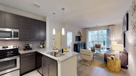 Fifth Street Place is a thoughtfully designed single-floor residence that seamlessly blends modern aesthetics with functional living. The open-concept kitchen serves as the heart of the home, featuring rich dark cabinetry with a sleek finish, a classic subway tile backsplash, and a granite-topped island with an undermount sink. Pendant lighting above the island adds a refined touch, casting warm light over the space. The kitchen flows naturally into the living area, which is bathed in natural light from large windows and a sliding glass door that opens onto a private balcony overlooking lush, wooded surroundings. The living space offers a generous and airy feel, with durable wood-style flooring that runs throughout, tying the areas together cohesively. The balcony provides a serene outdoor retreat with views of mature trees, enhancing the overall sense of tranquility. A hallway from the main living area leads to additional private spaces, completing this well-appointed home. The overall ambiance is contemporary and welcoming, making Fifth Street Place a polished and comfortable living environment.