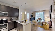 Fifth Street Place is a thoughtfully designed single-floor residence that seamlessly blends modern aesthetics with functional living. The open-concept kitchen serves as the heart of the home, featuring rich dark cabinetry with a sleek finish, a classic subway tile backsplash, and a granite-topped island with an undermount sink. Pendant lighting above the island adds a refined touch, casting warm light over the space. The kitchen flows naturally into the living area, which is bathed in natural light from large windows and a sliding glass door that opens onto a private balcony overlooking lush, wooded surroundings. The living space offers a generous and airy feel, with durable wood-style flooring that runs throughout, tying the areas together cohesively. The balcony provides a serene outdoor retreat with views of mature trees, enhancing the overall sense of tranquility. A hallway from the main living area leads to additional private spaces, completing this well-appointed home. The overall ambiance is contemporary and welcoming, making Fifth Street Place a polished and comfortable living environment.