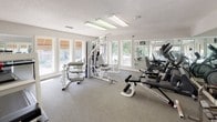This dedicated fitness center offers a comprehensive workout environment designed to support a wide range of exercise routines. The space is bright and welcoming, with multiple large windows along the walls that invite natural light and provide views of the surrounding greenery, creating an energizing atmosphere for exercise. Textured ceilings with recessed fluorescent lighting ensure the room is well-lit throughout.

The strength training area is well-appointed, featuring a multi-station cable machine suitable for a variety of resistance exercises, along with a dedicated free weight section complete with a full rack of dumbbells in graduated sizes and an adjustable weight bench. Additional benches and isolated strength machines round out the equipment offerings for targeted muscle training.

The cardio section is equipped with treadmills, elliptical trainers, and stationary cycles, all positioned to take advantage of the natural light. Full-length mirrors line one wall, visually expanding the space and allowing users to monitor their form during workouts. The flooring throughout is a durable, cushioned carpet well-suited for a fitness environment. Signage identifying the Fitness Center is prominently displayed, and a wall clock keeps users on schedule. Glass doors connect the fitness center to an adjoining common area, enhancing the sense of openness and accessibility.