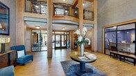 RiverWalk on the Falls presents a thoughtfully designed common area that blends rustic architectural elements with refined interior detailing. The grand lobby immediately establishes the tone of the space, featuring soaring double-height ceilings supported by substantial exposed timber columns. An interior mezzanine level with wrought-iron railings overlooks the main floor, adding vertical drama and architectural interest. A central round accent table with fresh floral arrangements anchors the entry area, which is finished with warm hardwood flooring and a patterned area rug.

Adjacent to the entry, a comfortable lounge area features a leather sofa, wingback seating, and a rustic wood coffee table beneath a statement circular chandelier. Large windows invite natural light and offer views of the surrounding landscape, while oversized abstract artwork adorns the textured gray walls.

A well-appointed corridor lined with reclaimed timber framing connects additional rooms, including glass-fronted private offices and meeting spaces. The consistent use of natural wood trim, warm neutral tones, and carefully chosen furnishings creates a cohesive and welcoming atmosphere throughout the entire facility.