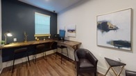 Located at 3920 Ivy Summit Ct in Cumming, Georgia, this single-floor professional office space combines functional design with a polished, contemporary aesthetic. The interior is defined by warm hardwood flooring that runs throughout, contrasting beautifully against crisp white walls and a bold navy accent wall that adds depth and character to the workspace.

The centerpiece of the office is a substantial L-shaped workstation crafted from reclaimed-style wood with sturdy bracket supports, offering generous surface area for computer equipment, printing devices, and daily workflow needs. High-back bar-style seating accompanies the workstation, creating an ergonomic and collaborative environment. Natural light filters in through a window fitted with white horizontal blinds, brightening the work area.

A dedicated seating nook near the French door entry provides a comfortable reception or consultation area, furnished with dark leather barrel chairs and a sleek accent table — ideal for client meetings or informal discussions. The French doors feature glass pane detailing and open the space to an adjacent area, enhancing the sense of connectivity.

Curated abstract artwork with navy, gold, and white tones is displayed throughout, reinforcing a cohesive and sophisticated color palette. Thoughtful accent lighting, including wall-mounted fixtures and a desk lamp, ensures the space remains well-lit and inviting. Overall, this office presents a refined, productive environment well-suited for professional use.