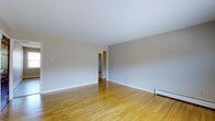 This single-floor space offers a straightforward and well-maintained layout designed for comfortable everyday living. The main living area is immediately welcoming, showcasing warm honey-toned hardwood floors that run throughout, adding a sense of continuity and warmth to the space. The walls are finished in a soft, neutral gray-white tone that reflects light evenly and provides a versatile backdrop for a variety of interior styles. White baseboards frame the perimeter of the room cleanly, complementing the overall minimal aesthetic. A doorway visible from the main room leads into an adjoining area, suggesting a connected and practical floor plan that allows for easy movement throughout the space. Baseboard heating units are positioned along the walls, providing an efficient and unobtrusive heating solution. The smooth, white ceiling contributes to an open and airy feel. The space is clean, neutral, and move-in ready, making it well-suited for those who value simplicity and functionality. Overall, the design prioritizes clean lines and a calm, understated atmosphere throughout.