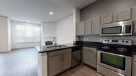 This single-floor unit presents a well-designed open-concept layout, with wood-style flooring extending consistently throughout the living spaces. The kitchen is equipped with stainless steel appliances, including a range, built-in microwave, and dishwasher, complemented by gray cabinetry with brushed metal hardware and dark countertops. A mosaic tile backsplash adds visual texture to the cooking area, while a peninsula with a double sink creates a natural division between the kitchen and the adjoining living space.

The living area is bright and generously proportioned, benefiting from large windows with blinds that allow ample natural light. A classically styled white fireplace serves as a focal point, flanked by French doors that provide additional light and outdoor access. Crown molding details and recessed lighting contribute to the polished atmosphere of the room.

A hallway connects the living areas to the private spaces of the unit, which include additional rooms with the same consistent flooring. The bathroom features a soaking tub with a fully tiled surround, accented by a distinctive black-and-white checkered tile border, adding a bold design element to the otherwise clean, neutral palette found throughout the space.