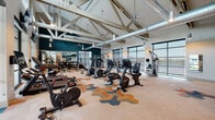 This well-appointed fitness center is defined by its striking architectural design, centered around soaring vaulted ceilings supported by exposed white-painted steel trusses. Industrial-style ductwork runs alongside the trusses, contributing to an open, loft-like aesthetic, while large ceiling fans and pendant lighting maintain a comfortable and well-lit environment throughout.

Floor-to-ceiling grid-frame windows line multiple walls, flooding the space with natural light and offering views of the surrounding exterior. A bold teal accent wall anchors the free weights area, providing visual contrast against the predominantly neutral palette. The flooring features a distinctive patterned carpet with geometric hexagonal shapes in earth tones, navy, and rust, adding a dynamic design element underfoot.

The fitness center is comprehensively equipped to support a full range of workouts. The cardio zone includes treadmills, elliptical trainers, stationary bikes, and stair climbers, many positioned to face the large windows. The strength area offers selectorized weight machines, a cable cross system, dumbbell racks ranging across multiple weights, medicine balls, resistance bands, and adjustable benches. Full-length mirrors in the free weights zone enhance spatial perception and support proper form during training sessions.