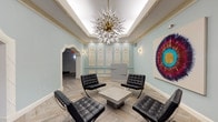 This sophisticated lobby space masterfully combines classical architectural elements with modern design sensibilities. The interior is defined by its soft sage-green walls, which provide a refined backdrop to a series of intricately detailed white plaster relief panels mounted along one wall. These ornamental panels, featuring elaborate neoclassical motifs, serve as a striking focal point and lend a sense of historic grandeur to the space.

The ceiling draws immediate attention with its tray design and illuminated cove lighting, from which a dramatic sputnik-style chandelier descends, featuring a brass core and radiating crystal-tipped spikes that add a bold, contemporary contrast to the classical surroundings. Ornate gold-toned wall sconces complement the chandelier throughout.

Underfoot, light-toned herringbone-patterned flooring adds geometric visual interest and warmth. A seating arrangement of black tufted leather Barcelona chairs surrounds a low concrete coffee table, offering a modern and functional gathering area at the heart of the lobby.

Arched doorways with detailed white molding frame passages to adjacent corridors and elevators, while a sleek modern reception desk sits against the decorative wall panels. A large-scale circular abstract artwork in vibrant jewel tones adds a contemporary artistic statement, completing the space's elegant and layered aesthetic.