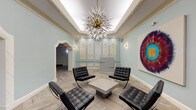 This sophisticated lobby space masterfully combines classical architectural elements with modern design sensibilities. The interior is defined by its soft sage-green walls, which provide a refined backdrop to a series of intricately detailed white plaster relief panels mounted along one wall. These ornamental panels, featuring elaborate neoclassical motifs, serve as a striking focal point and lend a sense of historic grandeur to the space.

The ceiling draws immediate attention with its tray design and illuminated cove lighting, from which a dramatic sputnik-style chandelier descends, featuring a brass core and radiating crystal-tipped spikes that add a bold, contemporary contrast to the classical surroundings. Ornate gold-toned wall sconces complement the chandelier throughout.

Underfoot, light-toned herringbone-patterned flooring adds geometric visual interest and warmth. A seating arrangement of black tufted leather Barcelona chairs surrounds a low concrete coffee table, offering a modern and functional gathering area at the heart of the lobby.

Arched doorways with detailed white molding frame passages to adjacent corridors and elevators, while a sleek modern reception desk sits against the decorative wall panels. A large-scale circular abstract artwork in vibrant jewel tones adds a contemporary artistic statement, completing the space's elegant and layered aesthetic.