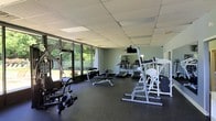 Located at 507 Sherwood Dr in Middletown, NY, this single-floor fitness facility offers a comprehensive workout environment designed to support a wide range of exercise routines. The gym is outfitted with an array of equipment including treadmills, a multi-function cable machine, free weight benches, a squat/power rack, and a dumbbell rack, making it suitable for both cardio and strength training needs. The space features durable rubber flooring throughout, providing safety and comfort during workouts. A drop ceiling with recessed fluorescent lighting ensures consistent, bright illumination across the entire floor. One of the standout features of this facility is the floor-to-ceiling glass wall along one side, which floods the interior with abundant natural light and offers pleasant views of the surrounding greenery and landscaping. A wall-mounted mirror near the strength equipment aids in form monitoring during training sessions. The sage-green accent walls contribute to a calm, focused atmosphere. The overall layout is open and spacious, allowing for comfortable movement between equipment stations and accommodating multiple users simultaneously.