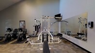 This fitness center is a dedicated single-floor exercise facility designed to meet a range of workout needs. The space is outfitted with a multi-station cable machine that enables a wide variety of strength training exercises, complemented by an adjustable weight bench for targeted muscle training. A rack of dumbbells lines one wall, offering options for free weight workouts of varying intensities. Additional cardio and resistance machines are positioned throughout the room, providing users with a comprehensive selection of equipment. The walls feature large mirrors that visually expand the space and allow users to monitor their form during exercise. Neutral-toned carpet tiles cover the floor, and the light gray walls contribute to a clean, focused atmosphere. A wall-mounted television is positioned above the equipment, allowing users to stay entertained during their workouts. Informational postings are displayed on the walls, offering guidance or safety instructions for equipment use. The overall layout is practical and efficiently organized, making good use of the available floor space while maintaining clear pathways between stations. The fitness center provides a functional and well-maintained environment suitable for a variety of exercise routines.
