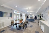Located in Chelsea, MA, this lobby and lounge offers a generously sized, single-floor layout that seamlessly blends welcoming functionality with contemporary design. The entrance greets visitors with wide sliding glass doors, light wood-look tile flooring, and tall modern planters that set an inviting tone from the moment of arrival. A reception desk sits prominently in the entryway, flanked by informational displays and a mounted television. Beyond the entry, the space transitions into a sophisticated lounge area anchored by a sleek marble-surround fireplace. Dramatic high-back navy accent chairs, a plush sectional sofa, layered area rugs, and curated artwork create a refined yet comfortable atmosphere. Recessed lighting and tray ceiling details add architectural depth throughout. A dedicated dining area features a striking live-edge wood table with upholstered seating, suspended crystal chandeliers overhead, and French doors that open directly to an outdoor patio, flooding the space with natural light. Glass-partitioned rooms provide semi-private lounge nooks with their own seating arrangements, enhanced by vibrant teal accent lighting. A staircase connects to additional levels, and thoughtful greenery throughout reinforces the space's fresh, modern character.