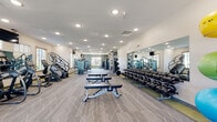 Located at 2210–2272 E Philadelphia St in Rapid City, South Dakota, this single-floor fitness center is designed to support a full spectrum of workout needs in a spacious, modern environment. The facility is thoughtfully organized into distinct training zones, allowing users to move seamlessly between cardio, strength, and functional training areas.

The cardio zone is outfitted with multiple treadmills, elliptical machines, a rowing machine, and a stair climber, providing a thorough range of aerobic options. Natural light filters in through generously sized windows framed with wood-toned accents, brightening the space and creating an inviting atmosphere. Recessed ceiling lighting supplements the natural light throughout.

The strength training area features an extensive dumbbell rack spanning a wide weight range, multiple weight machines targeting various muscle groups, a power rack, and flat benches. Full-length mirrors line the walls, enhancing the sense of openness while allowing users to monitor their form. A functional training station is equipped with resistance bands, TRX suspension trainers, stability balls, and additional accessories for varied training modalities.

Patterned, low-pile carpet tiles cover the floors throughout, lending comfort and durability underfoot. Mounted flat-screen televisions are positioned for easy viewing. A dual water fountain station is conveniently located near the entry, adjacent to glass doors leading to an adjoining common area. The overall ambiance is clean, modern, and well-maintained.