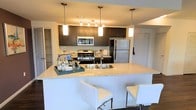 This single-floor residence showcases a thoughtfully designed open-concept layout that seamlessly blends style and functionality. The kitchen serves as a centerpiece of the space, featuring rich dark cabinetry that provides a striking contrast against light-toned countertops. Stainless steel appliances — including a gas range, built-in microwave, and full-size refrigerator — offer a polished, contemporary look. A generously sized kitchen island with a granite-style countertop doubles as a breakfast bar, complete with modern white bar stools, making it an ideal spot for casual dining or entertaining. Pendant lighting hangs elegantly above both the island and the kitchen workspace, while a track lighting system ensures the area is well-illuminated. The open design allows for a natural flow between the kitchen and adjoining living areas, creating a cohesive and inviting atmosphere. The neutral color palette throughout enhances the sense of openness, while clean lines and modern fixtures contribute to the overall contemporary ambiance. A glimpse of an adjacent room — possibly a closet or storage area — hints at additional practical features that complement this well-appointed living space.