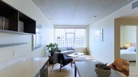 This thoughtfully designed single-floor urban residence offers a clean, contemporary aesthetic that makes excellent use of its layout. The living area serves as the heart of the space, anchored by large floor-to-ceiling windows that frame striking city skyline views and flood the interior with abundant natural light. The open design creates a seamless flow between the living and dining zones, with light hardwood flooring running throughout to enhance the sense of warmth and continuity. The ceiling features a refined paneled finish that adds a subtle architectural detail overhead. The living area is furnished with a minimalist sensibility — a streamlined sofa, a mid-century modern accent chair, and a sleek coffee table are arranged to invite both relaxation and conversation. Lush indoor plants introduce a natural, organic contrast against the predominantly white walls and neutral palette. A wall-mounted television and built-in cabinetry along one side provide functional storage while maintaining the space's uncluttered character. The kitchen area integrates smoothly into the open plan, featuring clean white cabinetry and ample counter space. Overall, the space exudes a calm, airy ambiance with a distinctly urban sensibility, making it well-suited for modern city living.