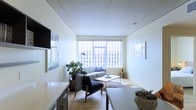 This thoughtfully designed single-floor urban residence offers a clean, contemporary aesthetic that makes excellent use of its layout. The living area serves as the heart of the space, anchored by large floor-to-ceiling windows that frame striking city skyline views and flood the interior with abundant natural light. The open design creates a seamless flow between the living and dining zones, with light hardwood flooring running throughout to enhance the sense of warmth and continuity. The ceiling features a refined paneled finish that adds a subtle architectural detail overhead. The living area is furnished with a minimalist sensibility — a streamlined sofa, a mid-century modern accent chair, and a sleek coffee table are arranged to invite both relaxation and conversation. Lush indoor plants introduce a natural, organic contrast against the predominantly white walls and neutral palette. A wall-mounted television and built-in cabinetry along one side provide functional storage while maintaining the space's uncluttered character. The kitchen area integrates smoothly into the open plan, featuring clean white cabinetry and ample counter space. Overall, the space exudes a calm, airy ambiance with a distinctly urban sensibility, making it well-suited for modern city living.