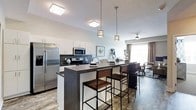 This single-floor space showcases a thoughtfully designed open-concept layout that seamlessly connects the kitchen, dining, and living areas. The kitchen is outfitted with stainless steel appliances, including a full-size refrigerator and built-in microwave, complemented by sleek white cabinetry and a dark mosaic tile backsplash that adds visual contrast. A spacious dark-topped kitchen island with a waterfall edge serves as the centerpiece, accommodating multiple bar-height stools with wood and metal frames, making it ideal for casual dining or entertaining. Overhead, a series of geometric gold-accented pendant lights hang above the island, adding a modern, upscale touch alongside flush-mount ceiling fixtures. The open living area beyond features a ceiling fan and access to natural light through large vertical window treatments. The flooring transitions between wood-look planks in the kitchen zone and a patterned area rug in the living space, defining each zone while maintaining a cohesive aesthetic. The overall ambiance is contemporary and well-appointed, balancing functional design with refined decorative details.