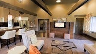 This single-floor space blends modern rustic design with a warm, welcoming ambiance. The main lounge area immediately draws attention with its soaring ceilings accented by bold, dark exposed beams that contrast beautifully against the warm tan walls. A prominent wood-paneled feature wall serves as a focal point, anchoring a wall-mounted flat-screen television and flanked by upholstered ottomans in coordinating coral and neutral tones. Comfortable seating, including a classic wingback chair adorned with accent pillows, is arranged around a sleek glass-topped coffee table, creating an inviting gathering area. The flooring transitions between rich dark hardwood and a neutral area rug, adding texture and warmth throughout the space. Adjacent areas visible from the main room suggest additional functional spaces, including what appears to be an office or reception area with a mirrored wall and organized shelving, as well as a secondary workspace. Decorative wall clocks and thoughtful accent pieces contribute to the space's curated, lived-in character. Recessed lighting provides consistent illumination throughout, complementing the overall cozy yet polished aesthetic of this well-appointed communal environment.