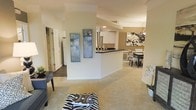 This single-floor residence showcases a thoughtfully designed open-concept layout that seamlessly connects the living, kitchen, and dining areas. The living space features plush carpeting in warm neutral tones, complemented by tasteful décor including bold abstract artwork and accent pieces that add personality to the room. A central structural column with a half-wall divides the space while maintaining an airy, open feel throughout.

The kitchen is positioned at the heart of the home, featuring a peninsula with a dark countertop that serves as both a functional workspace and a casual gathering spot. White cabinetry lines the kitchen walls, providing ample storage, while recessed lighting illuminates the area effectively.

Adjacent to the kitchen, the dining area offers a dedicated space for meals, furnished with a classic dining table and patterned chairs that add a touch of visual interest. Decorative wall art enhances the ambiance of this corner of the home.

Hardwood-style flooring in the entryway transitions to carpeted areas in the main living zones, creating a natural definition between spaces. Doors visible along one corridor suggest additional rooms, including storage and utility areas. The overall ambiance is warm, inviting, and move-in ready, with cohesive design elements tying each area together harmoniously.
