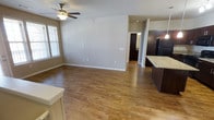 This single-floor space offers an open-concept layout that seamlessly connects the living area and kitchen, creating a sense of spaciousness and flow. The living area is finished with warm hardwood-style flooring that extends throughout, complementing the neutral gray-toned walls and white baseboards. A ceiling fan with a built-in light fixture is centrally mounted, providing both airflow and ambient lighting. Natural light filters into the space through a window dressed with horizontal blinds, brightening the room and highlighting the clean, understated finishes. Multiple electrical outlets are positioned along the walls, offering practical convenience throughout the living space.

The kitchen occupies one side of the open floor plan and features rich, dark espresso-toned cabinetry paired with light granite countertops, creating a striking visual contrast. A black refrigerator anchors the kitchen, and the cabinetry appears to extend generously for storage. A doorway visible toward the rear of the space leads to additional rooms, suggesting a functional layout that balances communal and private areas. Overall, the space presents a modern, clean aesthetic with cohesive finishes throughout.