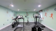 Located at 1053 Rifle Range Rd in Mt. Pleasant, South Carolina, this single-story fitness center offers a well-equipped and inviting environment for exercise and wellness. The space is finished with soft mint-green walls accented by decorative wall art, creating a lively and motivating atmosphere. Recessed lighting throughout the ceiling ensures bright, even illumination, while a ceiling fan provides comfortable airflow during workouts. The floor is covered in durable, patterned commercial carpet, adding both comfort and practicality underfoot. The cardio equipment area is well-appointed, featuring treadmills, an elliptical trainer, a stationary upright bike, and additional exercise machines, providing a variety of options for cardiovascular fitness. A window along one wall allows for natural light to filter into the space, enhancing the overall ambiance. Framed motivational artwork and decorative accents on the walls contribute to an encouraging and pleasant workout environment. The layout is thoughtfully organized to allow users to move comfortably between equipment. This fitness center delivers a functional and welcoming space suitable for a range of fitness routines and activity levels.