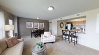 Located at 6216 66th St in Lubbock, Texas, this single-floor residence offers a thoughtfully designed open-concept floor plan that seamlessly connects the living, dining, and kitchen areas into one cohesive and functional space. The living area is finished with plush wall-to-wall carpeting in a warm neutral tone, creating a comfortable and inviting atmosphere. Taupe-painted walls provide a clean, modern backdrop that complements the natural wood and soft furnishings throughout. A dedicated dining area sits adjacent to the main living space and features ample room for a full dining set, accented by a triptych of framed artwork on the wall. The kitchen flows naturally from the dining area and is equipped with wooden cabinetry, a full-size refrigerator, and a raised breakfast bar with seating — ideal for casual dining or entertaining. Overhead lighting is flush-mounted throughout, maintaining clean sight lines across the open layout. HVAC vents are integrated into the ceiling for efficient climate control. A doorway visible from the main living area leads to additional private spaces within the unit. The overall design balances practicality with a warm, residential aesthetic, making this a comfortable and well-appointed single-level home.