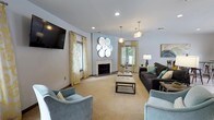 This single-story clubhouse offers a thoughtfully designed communal lounge space that balances comfort and contemporary style. The main gathering area is anchored by a classic fireplace framed by a clean white mantel, serving as a natural focal point for the room. Soft blue-gray walls create a calm, inviting atmosphere, complemented by neutral carpeting that adds warmth underfoot.

Natural light filters in through multiple windows dressed in gold and cream patterned curtains, lending the space a bright and airy feel. Overhead, a combination of recessed lighting and stylish pendant fixtures with a warm metallic finish provides versatile illumination throughout the day and evening.

The seating arrangement is carefully curated, featuring a large dark charcoal sofa accented with teal and patterned throw pillows, alongside plush powder-blue accent chairs that invite relaxed conversation. A sleek rectangular coffee table with a marble-style top ties the seating area together. A wall-mounted flat-screen television adds a functional entertainment element, while a distinctive circular metallic wall sculpture adds an artistic finishing touch to the overall design.