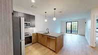 Located at 199 Gardner Street in Boston's West Roxbury neighborhood, this thoughtfully designed one-bedroom, two-bathroom unit offers a modern open-concept layout across a single floor. Light wood-tone flooring flows throughout the space, complementing the clean white walls and contemporary black hardware accents found on doors and fixtures.

The combined kitchen and living area serves as the heart of the unit. The kitchen is appointed with two-tone cabinetry — warm wood lower cabinets paired with charcoal upper cabinets — along with quartz countertops, a stone tile backsplash, stainless steel appliances, and a peninsula with pendant lighting that provides additional prep and seating space. The living area is bright and open, anchored by floor-to-ceiling sliding glass doors that lead to a private balcony overlooking a canopy of mature trees, bringing in generous natural light and a serene green outlook.

The bedroom is a comfortable, well-proportioned space featuring a double-door closet and the same warm flooring found throughout. Both bathrooms are finished in a clean, minimalist style with light tile flooring, wood-accented vanities, white countertops, and frameless glass shower enclosures with matte black hardware. A hallway connects the rooms with efficient flow, completing this well-appointed Boston residence.