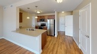 This single-floor residence offers a thoughtfully designed layout with a variety of functional spaces finished with quality materials throughout. The open-concept kitchen serves as a central hub, featuring warm wood cabinetry, granite-style countertops, a kitchen island with a built-in sink, and a full suite of stainless steel appliances including a refrigerator, electric range, and microwave. Pendant lighting above the island adds a polished touch, while hardwood-style flooring extends seamlessly into the adjoining living and dining areas. The living area is bright and spacious, bathed in natural light from multiple windows dressed with horizontal blinds and a sliding glass door fitted with vertical blinds that opens to an outdoor view. A pendant light fixture anchors the dining space, which connects to the rest of the unit with ease. The bedrooms feature plush wall-to-wall carpeting, generous natural light from large windows, and ceiling fans for added comfort. The full bathroom is well-equipped with a wood-vanity featuring a granite-style countertop, a walk-in shower with glass door, a safety grab bar, and warm-toned tile flooring. Neutral beige walls and white trim carry a clean, cohesive aesthetic throughout the entire space.