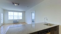 This well-designed single-floor residence offers a seamless open-concept layout that connects the kitchen, dining, and living areas into one cohesive and functional space. The kitchen is anchored by a generous granite countertop island with a sleek, modern faucet and sink, complemented by a dishwasher and contemporary track lighting overhead. The light-toned granite surface adds both elegance and practicality to the space.

The living area features warm hardwood flooring that extends throughout, enhancing the brightness already provided by a large multi-pane window. This window allows abundant natural light to flood the interior, creating an airy and inviting atmosphere. A wall-mounted heating unit beneath the window ensures year-round comfort.

The clean white walls throughout the unit provide a neutral, versatile backdrop suited to a variety of interior styles. A doorway visible from the main living area leads into a private bedroom, which also benefits from natural light through its own window with vertical blinds. The overall design is characterized by simplicity and functionality, with an emphasis on open sightlines and a bright, modern aesthetic that makes the space feel welcoming and comfortable.