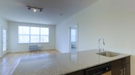This well-designed single-floor residence offers a seamless open-concept layout that connects the kitchen, dining, and living areas into one cohesive and functional space. The kitchen is anchored by a generous granite countertop island with a sleek, modern faucet and sink, complemented by a dishwasher and contemporary track lighting overhead. The light-toned granite surface adds both elegance and practicality to the space.

The living area features warm hardwood flooring that extends throughout, enhancing the brightness already provided by a large multi-pane window. This window allows abundant natural light to flood the interior, creating an airy and inviting atmosphere. A wall-mounted heating unit beneath the window ensures year-round comfort.

The clean white walls throughout the unit provide a neutral, versatile backdrop suited to a variety of interior styles. A doorway visible from the main living area leads into a private bedroom, which also benefits from natural light through its own window with vertical blinds. The overall design is characterized by simplicity and functionality, with an emphasis on open sightlines and a bright, modern aesthetic that makes the space feel welcoming and comfortable.