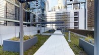 This outdoor courtyard is a well-designed urban retreat set within a dense city environment, surrounded by modern glass and concrete high-rise buildings that frame the space on multiple sides. A wide, light-colored paved walkway runs centrally through the courtyard, providing a clean, unobstructed path through the area. Overhead, sleek metal pergola structures line the walkway, offering partial shade and adding an architectural element that complements the contemporary surroundings. The courtyard features carefully maintained green spaces, including patches of low ground cover and young trees that introduce a natural contrast to the hard urban landscape. Raised rectangular planters with dark charcoal finishes are positioned along the perimeter, contributing to the greenery and helping define the boundaries of the space. Bollard-style lighting is placed along the walkway, suggesting usability during evening hours. Trash receptacles and additional seating or utility fixtures are visible throughout, indicating the space is designed for regular use. The overall ambiance is modern and functional, offering a calm, landscaped passage or gathering area within an otherwise dense urban setting.